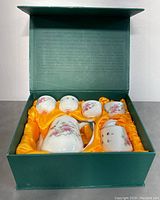Open box view showing porcelain teapot, creamer, sugar bowl and four tea cups nestled in yellow satin lining.
