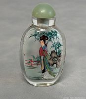 Front view of the snuff bottle showing a reverse painted Chinese woman in red, blue, and green robes by bamboo and rock landscape, green stopper on top.