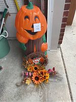 Large plastic pumpkin figure with smiling jack-o'-lantern face, autumn wreath, and floral arrangement displayed on a concrete floor next to a brick wall