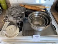 Photo showing two wooden rolling pins, stack of 3 stainless steel mixing bowls inside a large metal baking pan, kitchen scale with measuring cup on top, and several metal baking trays stacked next to them along with wire racks.