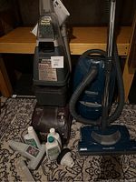 Kenmore vacuum and Hoover SteamVac carpet shampoo machines with hoses and wands on floor next to wooden shelving