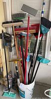 Various upright brooms, mops, dust pans, and a cordless Hoover vacuum leaning in a bucket