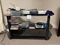 Glass 3-tier TV stand holding various audio and video components including stacks of CDs, cassette tapes, remote controls, cords, and a blue and black Bluetooth headset.