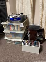 Curtis radio and Electrohome VHS player on top of plastic bins containing VHS tapes, with wooden and white CD racks next to them