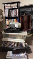 Photo showing entire lot with stacked media racks including CDs, DVDs, VHS tapes, a Sharp VHS player, Sony radio CD player, headphones, and a wooden DVD shelf.