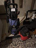 Photo of blue Simonz 2000PSI power washer with hose reel and black handle, alongside a red and black Craftsman 1.6 gallon wet/dry vacuum with attached hose, placed on patterned carpet in basement.