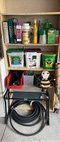 Three shelves with gardening books, plant foods, chemicals, gardening pouch, hose heads, and various containers. Gardening bench below with coiled black hose and bee garden stake decoration nearby.