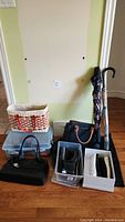 Photo showing two umbrellas leaning against the wall next to multiple handbags including a woven basket-style handbag, a black handbag, and a dark blue handbag with tan handles. Also visible are two plastic bins containing shoes and other accessories.