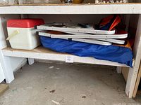 Folded two lawn chairs stacked on blue carrying bags under a shelf with white legs and gray concrete floor.