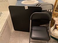 Photo showing folded black square table along with stacked black folding chairs in a tiled room.