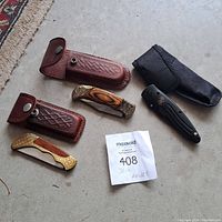 Image showing three folding pocket knives and two brown leather sheaths with snap closures placed on concrete floor alongside a black fabric pouch and a MaxSold lot ticket.