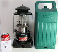 Front view of Coleman double mantle lantern with glass globe and protective case side by side, includes size reference Coke can and lot number card.