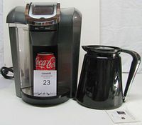 Front view of Keurig 2.0 coffee maker with black 4 cup carafe beside it; a standard Coca-Cola can placed inside coffee maker's brew area for scale.