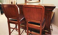 View of two wooden bar stools placed side by side in front of the solid wood bar counter, showing the backrest design and padded seats
