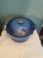 Blue large cast iron pot with lid and metal knob stamped with Wolfgang Puck logo, showing side handles.