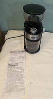 Black & Decker branded coffee grinder with attached power cord and safety instructions sheet laid next to it on a white surface.