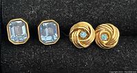 Two sets of blue earrings placed on dark fabric background. One pair has rectangular light blue faceted stones in gold-tone setting. The other pair shows gold-tone swirl design with small round light blue stones in center.