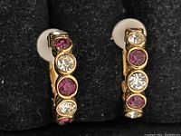 Close-up image of a pair of gold tone hoop earrings with alternating clear and pink stones set in bezels along the front.