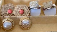 Photo shows three pairs of clip-on earrings in a jewelry box: hexagonal carved with coral-red center, rectangular faux pearl opalescent, and round gold-tone with large faux pearls.