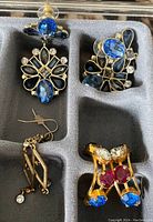 Photo showing three pairs of earrings in a foam storage case: blue rhinestone drops, gold geometric dangles, and gold with colorful round rhinestones.