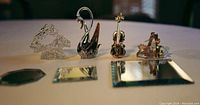 Four glass and Swarovski figurines displayed on mirrored tiles: clear glass swan, brown Swarovski crystal swan, crystal violin with bow, and rocking horse figurine.