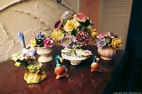 Porcelain floral and bird figurines arranged on a wooden surface displaying bouquets in vases, flower baskets with lace accents, and colorful ducks.