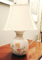 Clear photo of the lamp on a wooden surface showing the ceramic base with hand-painted colorful flowers and the white fabric lampshade.