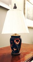 Bedside table lamp with dark blue ceramic vase-style base decorated with red flowers and a white pleated fabric lampshade with decorative finial.