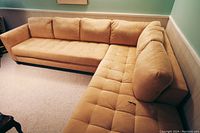 L-shaped tan sectional sofa with tufted seat cushions and loose back cushions against green wall
