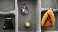 Image showing four necklace pendants in gray jewelry organizer tray: a black trapezoid pendant, a pink faceted oval pendant, a small geometric gold-tone pendant, and a large amber-colored triangular pendant with dark inclusions.