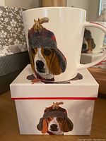 Photo of a white ceramic coffee mug featuring a beagle dog wearing a red and gray trapper hat with a squirrel on top, placed on top of matching box showing the same design.