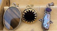 Three necklace pendants displayed together in a beige jewelry box. Left pendant is oval polished brown stone, center pendant round black sunburst with gold border, right pendant abalone shell and amethyst cabochon in silver-tone setting.