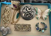 Seven assorted pins laid out on a cream-colored fabric background inside a blue box, showing various designs including abstract shapes, floral details, butterfly, horseshoe, and a dog pin.