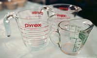 Three vintage Pyrex glass measuring cups, two 2-cup cups with red metric markings and one 1-cup with green markings.