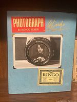 Cover of the Photograph book by Ringo Starr showing a camera lens with Ringo's photo and a label referencing photographic prints.
