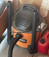 Photo shows a Rigid brand orange and black wet/dry shop vacuum with attached hose and two upright extension tubes beside it.