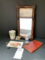Photo showing the small wooden framed wall mirror with sepia print inset, the Limoges cup with floral design, the Daum Nancy cameo glass cordial, two small shot glasses, a small spoon, an antique postcard, and the leather bound almanac.