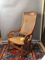 Front angled view of antique wooden rocking chair showing caned seat and back, curved armrests with scroll design, and wood grain.