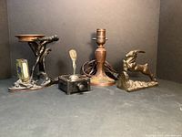 Set of four Art Deco decorative items shown against a dark neutral background, including ash tray, lighter, lamp base, and oryx bookend.