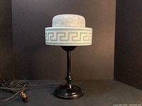 Full view of the table lamp showing frosted glass shade with Greek key and floral acid etching, black metal base, and attached electrical cord.
