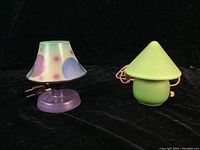 Set of two bedside lamps on black background. Left lamp has colorful pastel-spotted conical shade and purple base. Right lamp is green with conical shade and Greek key pattern.