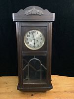 Front view of the wooden antique Divina wall clock showing the carved top, clock face with black Arabic numerals, brass bezel, and glass door panels protecting the face and pendulum.