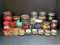 Full view of all 23 glass jars filled mostly with buttons sorted by color including red, white, green, black, blue, brown, beige, and multicolored. Jar types include vintage cosmetic jars, Ball mason jars, and pickle jars.