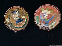 Photo of both terracotta plates on stands against black background showing Day of the Dead skeletal figures with musical and fruit basket themes.