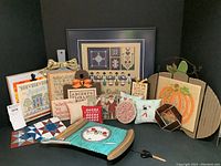 Full view of the framing cross stitch, decorative pillows, metalwork quilt squares, and clock sitting on cloth backdrop