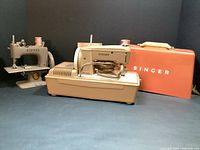 Singer Simanco Sewhandy Model 20 metal sewing machine on the left, Singer Sewhandy electric sewing machine on the right with orange carrying case behind it, all on dark background.