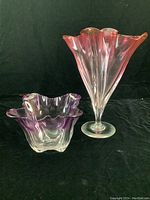 Front view of both pieces on black backdrop: purple-topped ruffled bowl at left, ruby-topped fluted vase at right