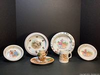 Seven pieces of vintage German nursery ware, including a creamer, underplate, two baby plates, two baby dishes, and a small cup with colorful nursery-themed hand-painted designs.