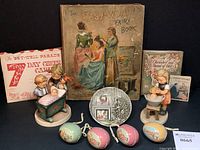 Photo showing the assortment of items including the two M.I. Hummel figurines flanking decorative items, tin Easter eggs, Christmas saucer, books and cards arranged on a surface.