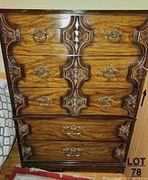 Front view showing 5 drawers with ornate wood carved trim and metal drawer pulls, wood grain finish and signs of wear.